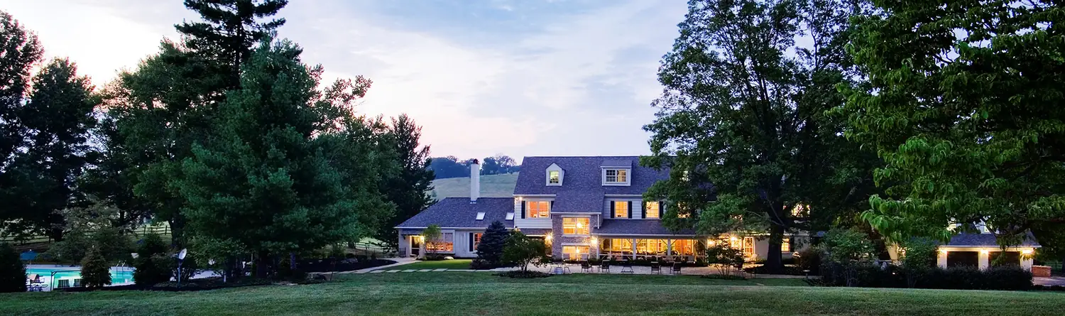 The estate house at dusk, its windows lit, seen across the lawn.